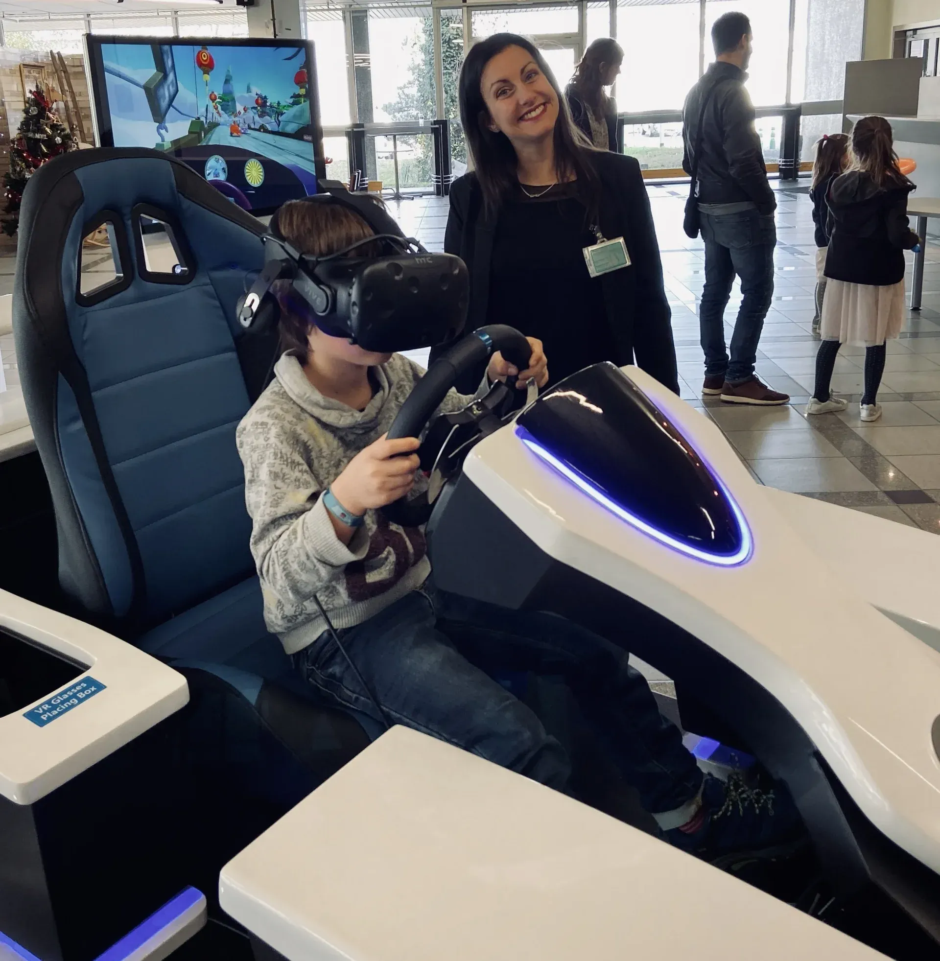 Enfant en casque VR au volant du simulateur kart Evos accompagné d'une animatrice souriante dans un hall vitré