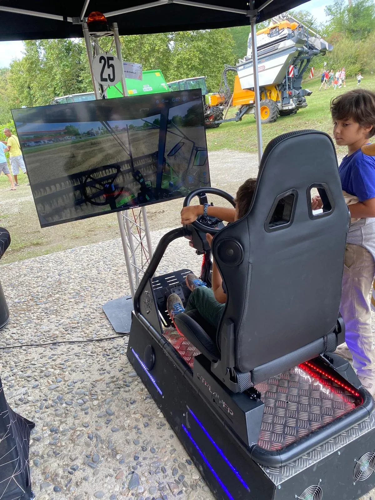Enfant aux commandes du simulateur agricole avec volant et écran sous un barnum lors d'une fête en plein air