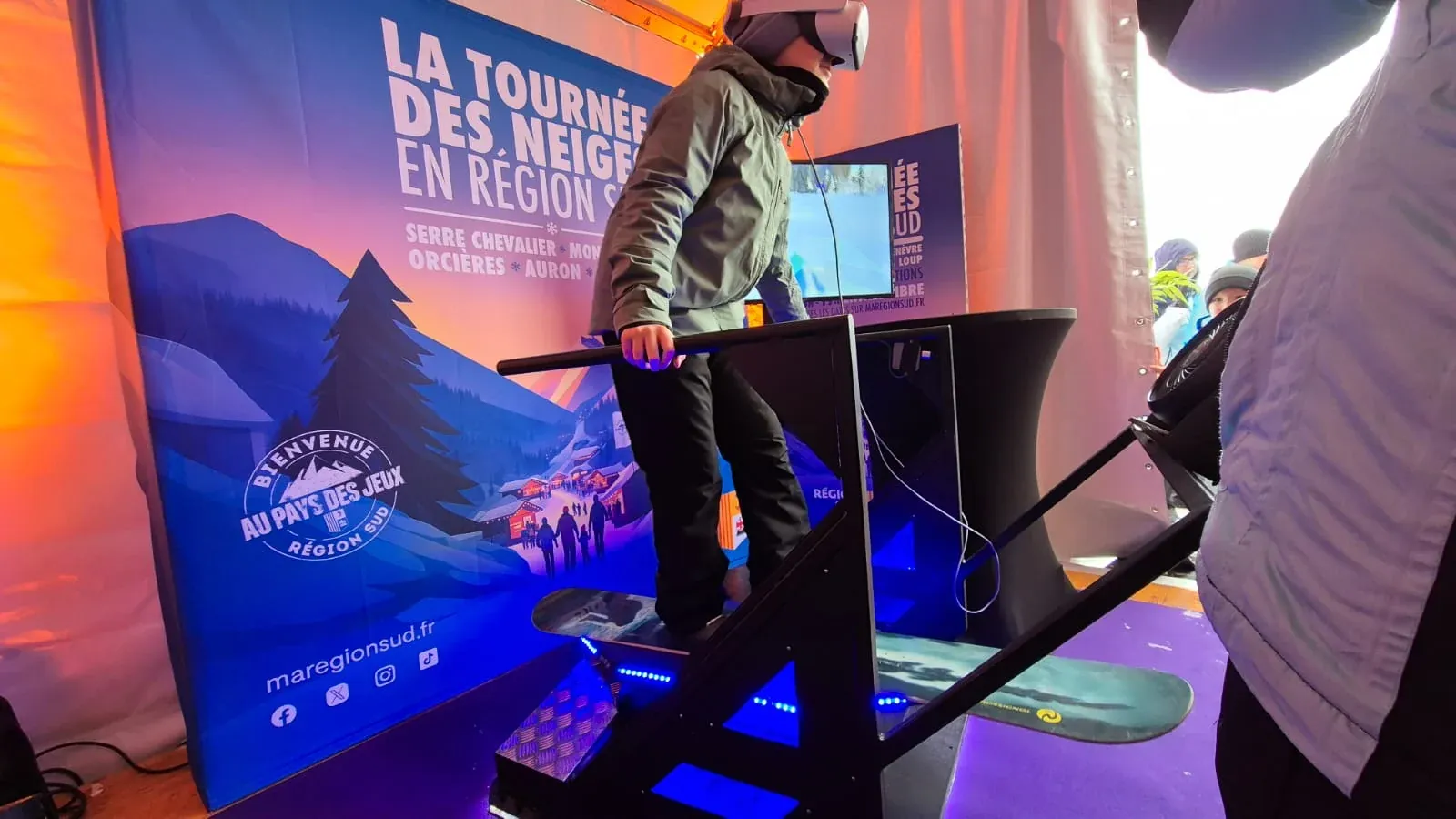 Jeune participant en sweat à capuche et casque VR sur un simulateur snowboard avec LED bleues devant une bâche Tournée des Neiges Région Sud