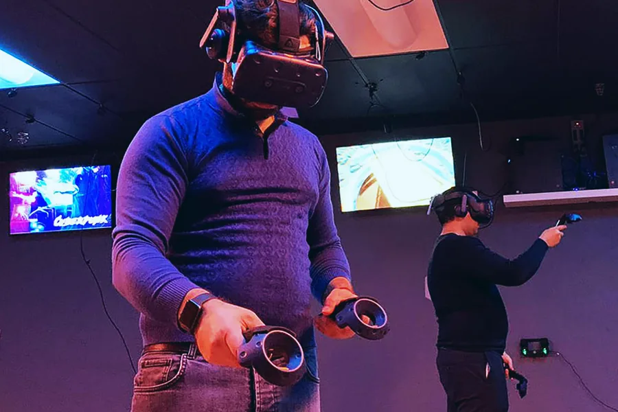 Joueur avec casque VR et deux manettes dans une salle sombre, second participant en arrière-plan