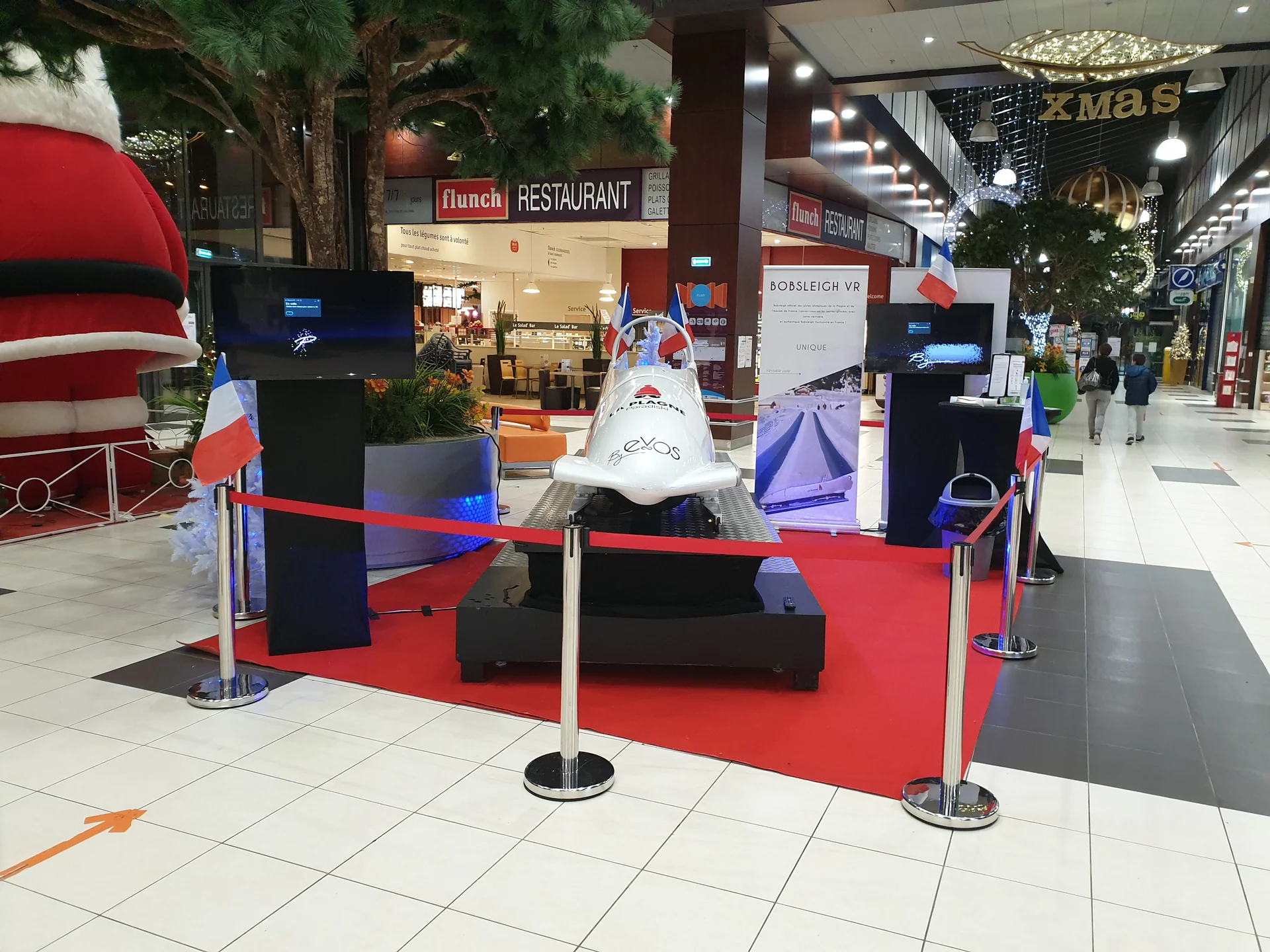 Vue d'ensemble du stand bobsleigh VR face au restaurant Flunch avec écrans de retransmission et drapeaux français