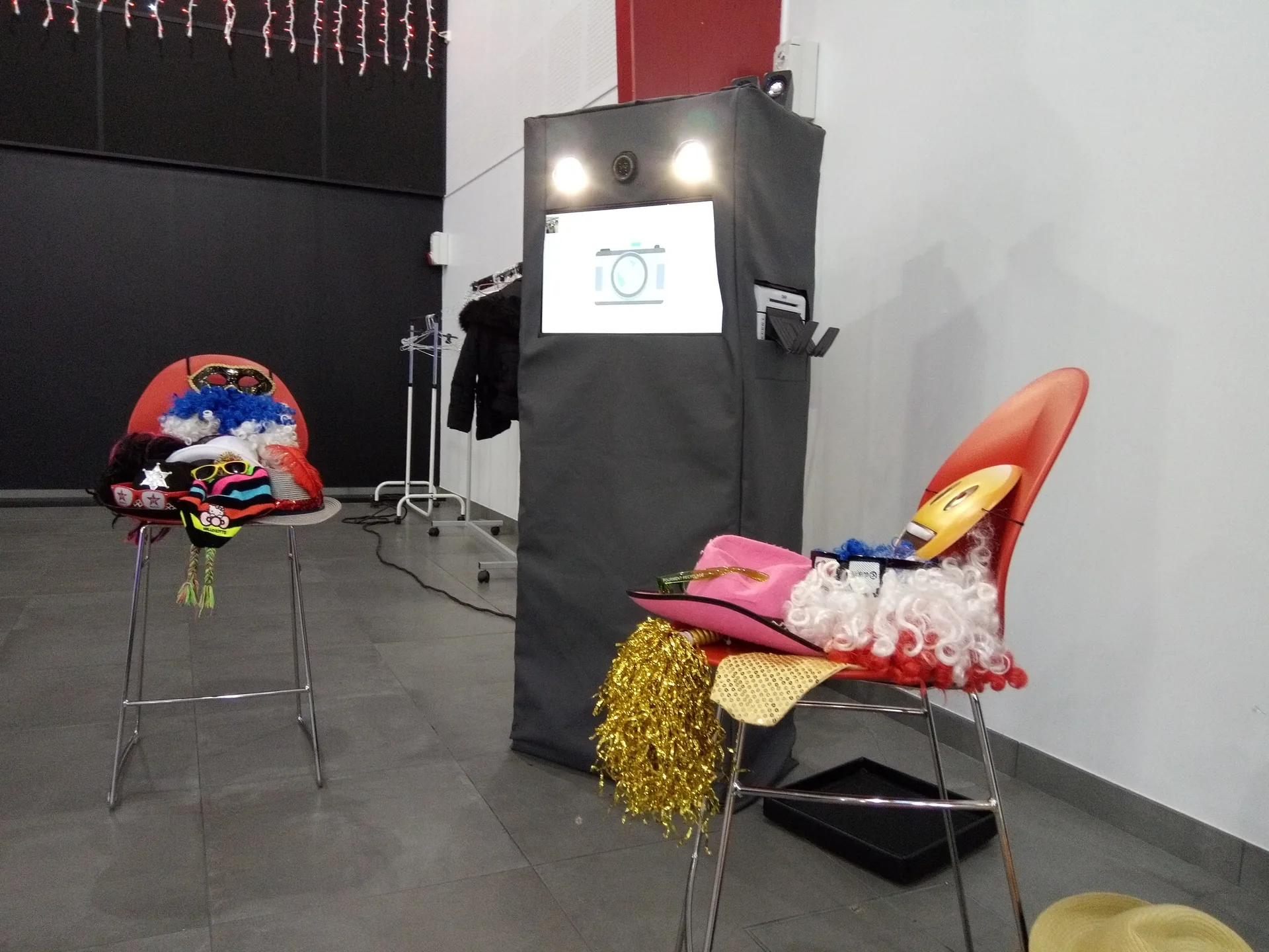 Borne photobooth avec perruques colorées et chapeaux fantaisie sur tabourets dans la salle des fêtes municipale