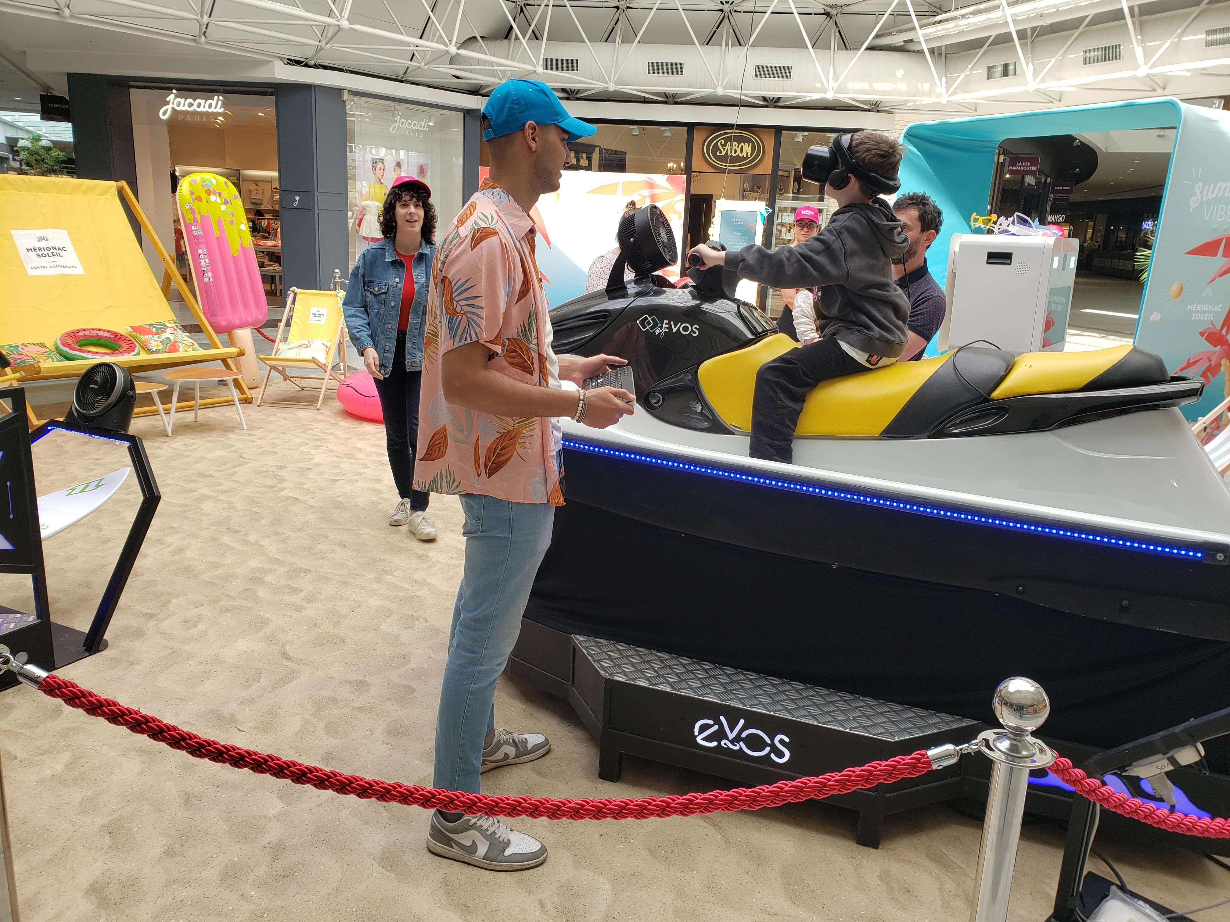 Animateur en chemise tropicale orange devant le jet ski VR balisé par un cordon rouge avec transats jaunes en arrière-plan