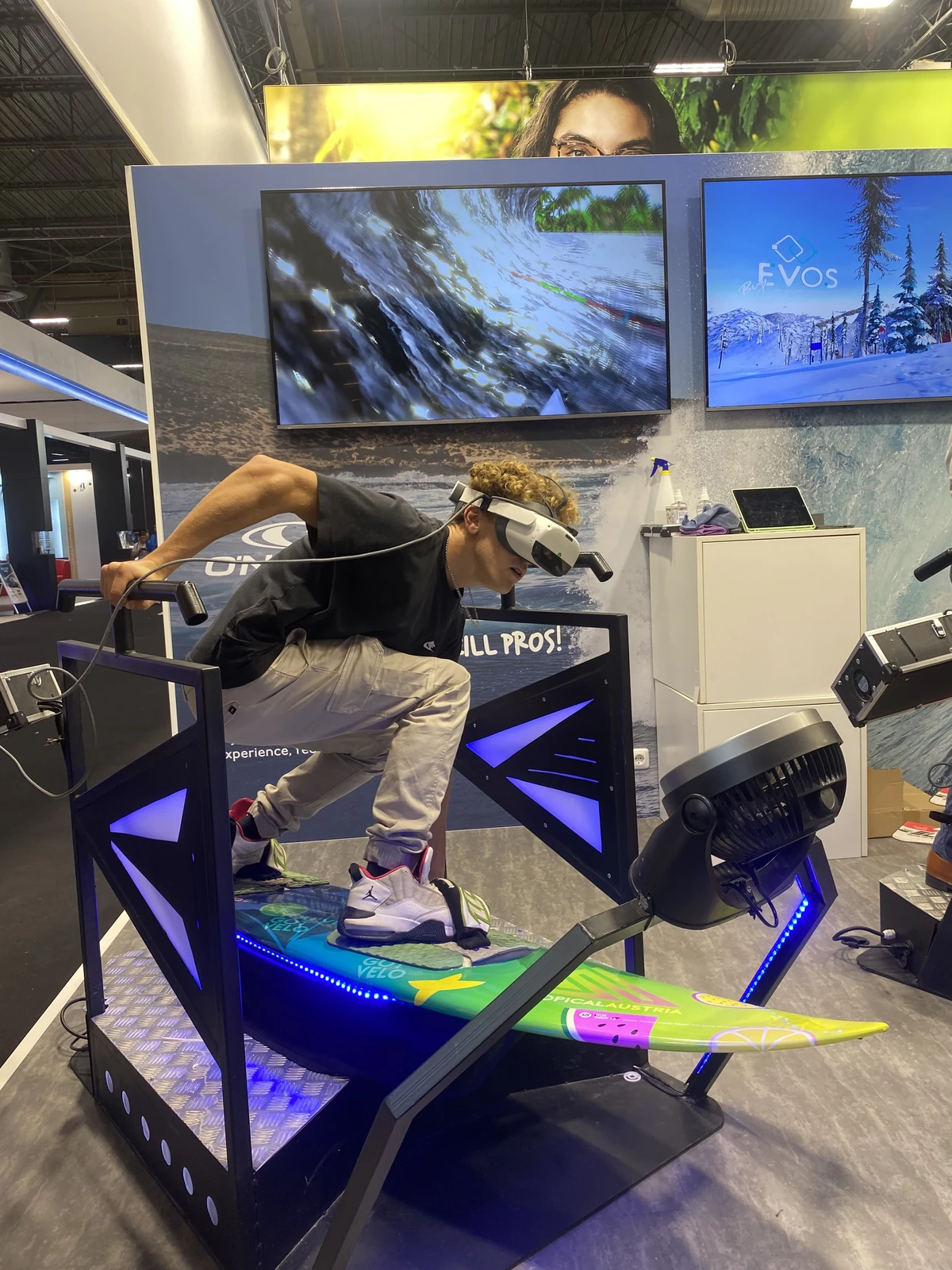 Participant cheveux bouclés sur simulateur surf VR avec écrans du stand Eschenbach en arrière-plan
