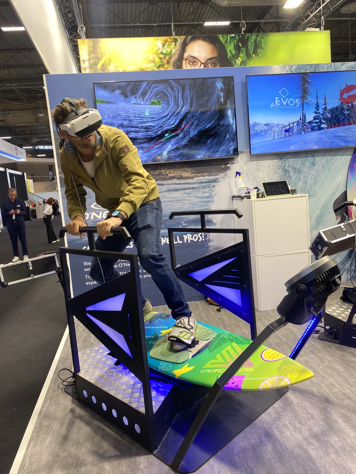 Visiteur en veste kaki sur le simulateur surf VR avec planche jaune et écran TV diffusant la session